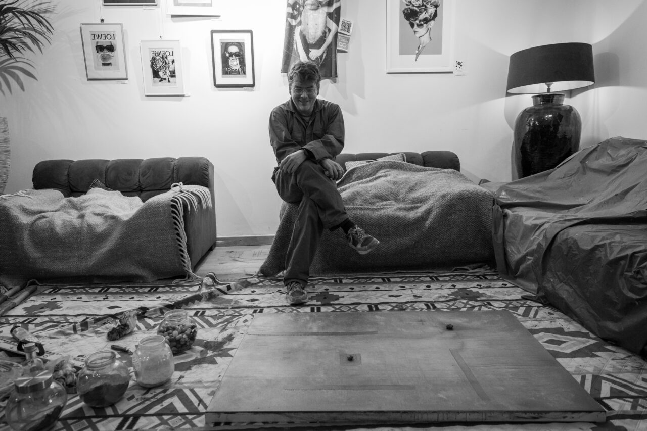 Black and white portrait of Ward Schepman seated on a sofa, with artworks on the wall.